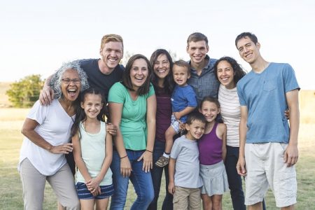 Portrait of an ethnically diverse, multi-generation family outdoors in warm weather. They are standing and smiling towards the camera.